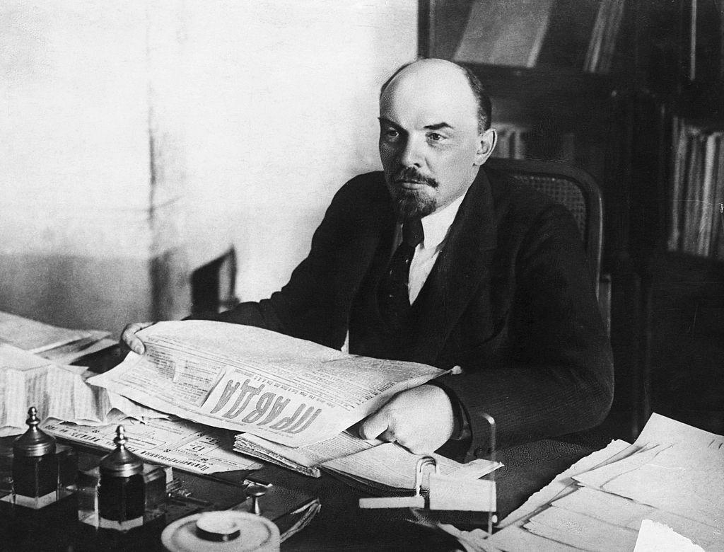 Foto en blanco y negro de Lenin sentado en su escritorio con un ejemplar del periódico Pravda en sus manos.