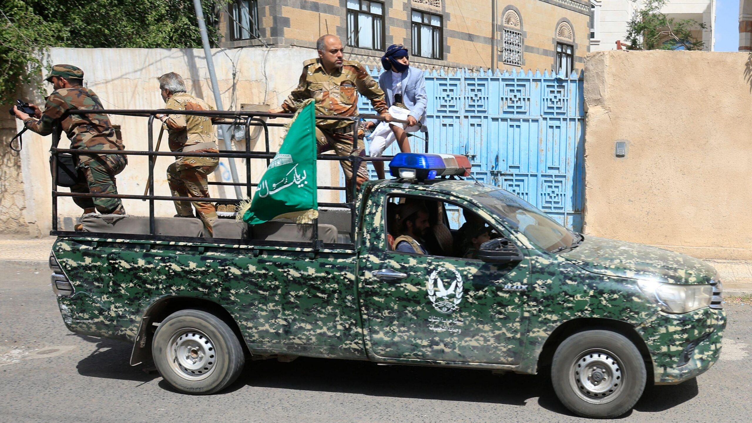 Cuatro hombres viajan en la parte trasera de una camioneta por una calle de Sana'a el 10 de septiembre de 2026. Uno de ellos sostiene una bandera verde sobre el lateral de la jaula metálica en la parte trasera del vehículo. Pasan junto a un edificio de ladrillo marrón claro con una puerta metálica azul claro.