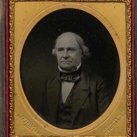 Una foto contemporánea en blanco y negro de un hombre maduro de fin de los 1800. Viste un saco y chaleco negro sobre una camisa blanca, un corbatín negro y cuello almidonado