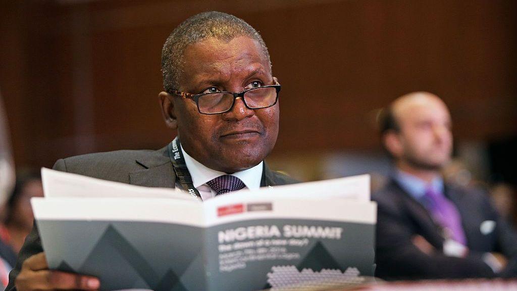 Dangote asiste a una conferencia y mira por encima de sus gafas. Sostiene un folleto titulado "Cumbre de Nigeria". Viste un traje gris, camisa blanca y corbata azul, y lleva un cordón alrededor del cuello.