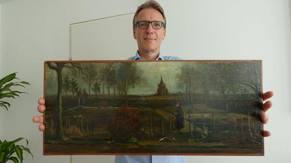 Un hombre con gafas y camisa azul sostiene un cuadro frente a la cámara. La imagen, bastante oscura, muestra una figura caminando por un jardín, con un edificio que parece una iglesia al fondo.