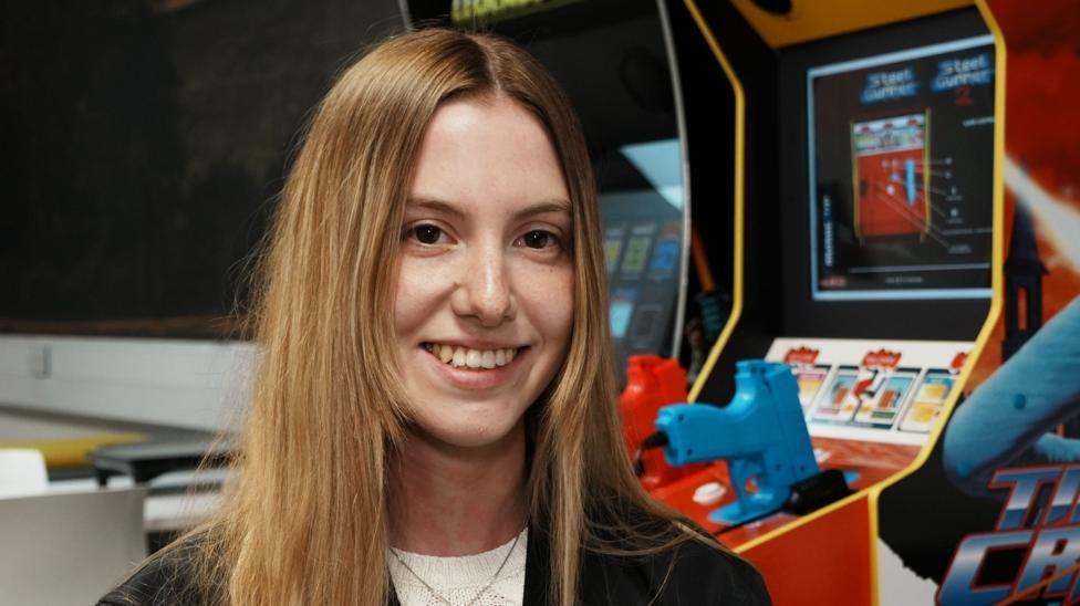 Mujer de cabello rubio sonríe a la cámara. Detrás de ella hay varios video juegos