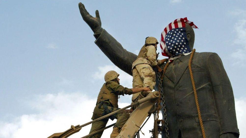 Tropas estadounidenses derriban una gran estatua de Saddam Hussein en Bagdad tras la invasión de Irak liderada por Estados Unidos en 2003, mientras una bandera estadounidense colocada sobre la cabeza de la estatua cubre su rostro.