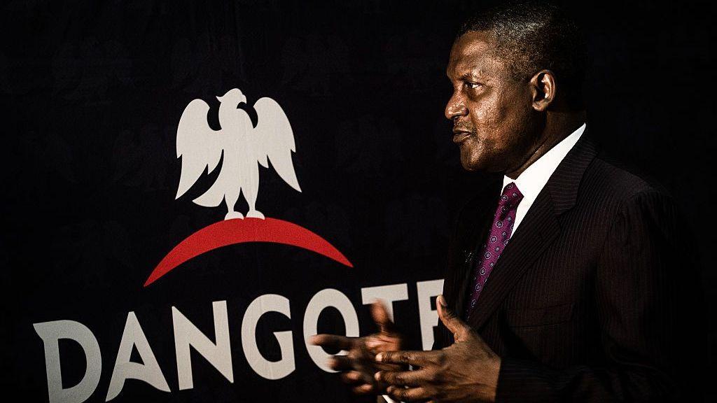 Dangote se encuentra frente al logotipo de su empresa, que consiste en su nombre en letras blancas en negrita y la silueta de un pájaro. Viste un traje negro, camisa blanca y corbata granate, y gesticula con las manos. 