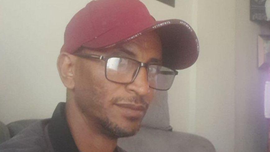 Un hombre de mediana edad, con gafas y gorra roja, está sentado en un sofá. 