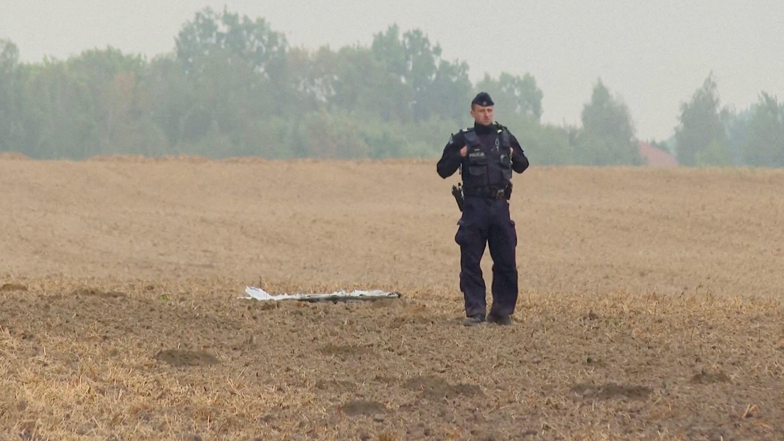 Un agente de policía se encuentra en un campo cosechado cerca de un trozo de escombro tirado en el suelo, con una hilera de árboles visible al fondo bajo un cielo brumoso.
