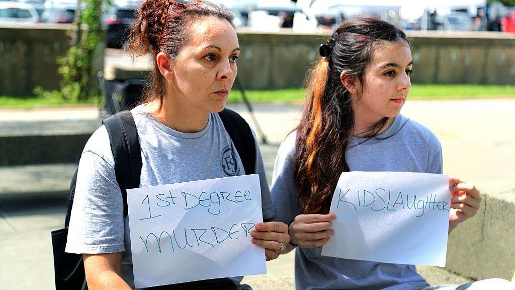 Se ve a dos personas con carteles fuera del juzgado. Los carteles dicen "Asesino en primer grado" y "Homicidio infantil".