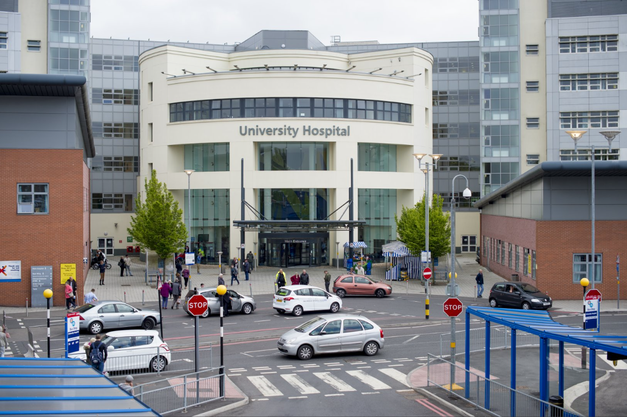 Exterior del Hospital Universitario de Coventry. Tiene una entrada semicircular con ventanas y una puerta ancha, flanqueada por edificios de ladrillo rojo.