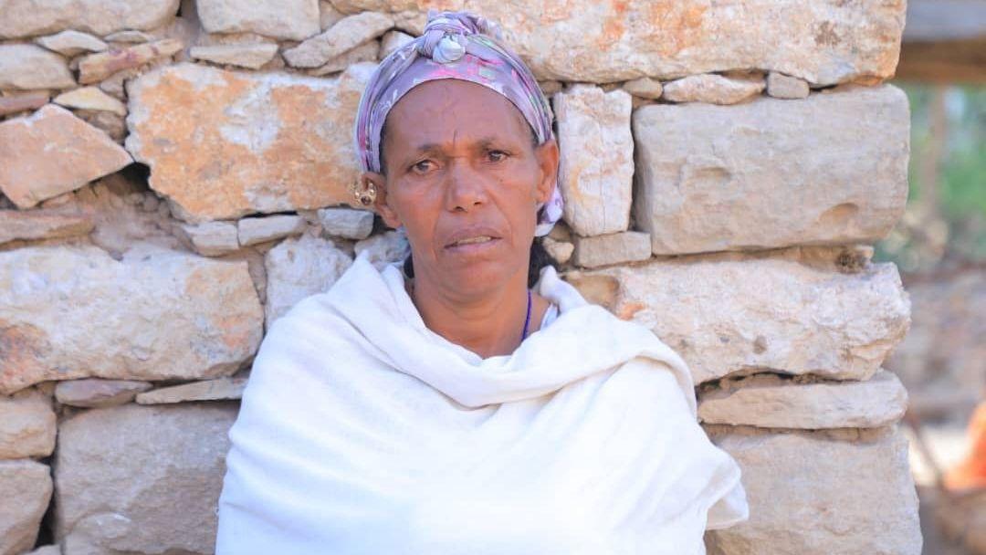 Una mujer está sentada, con la espalda apoyada en un muro de piedra. Lleva un chal blanco y un pañuelo morado en la cabeza.