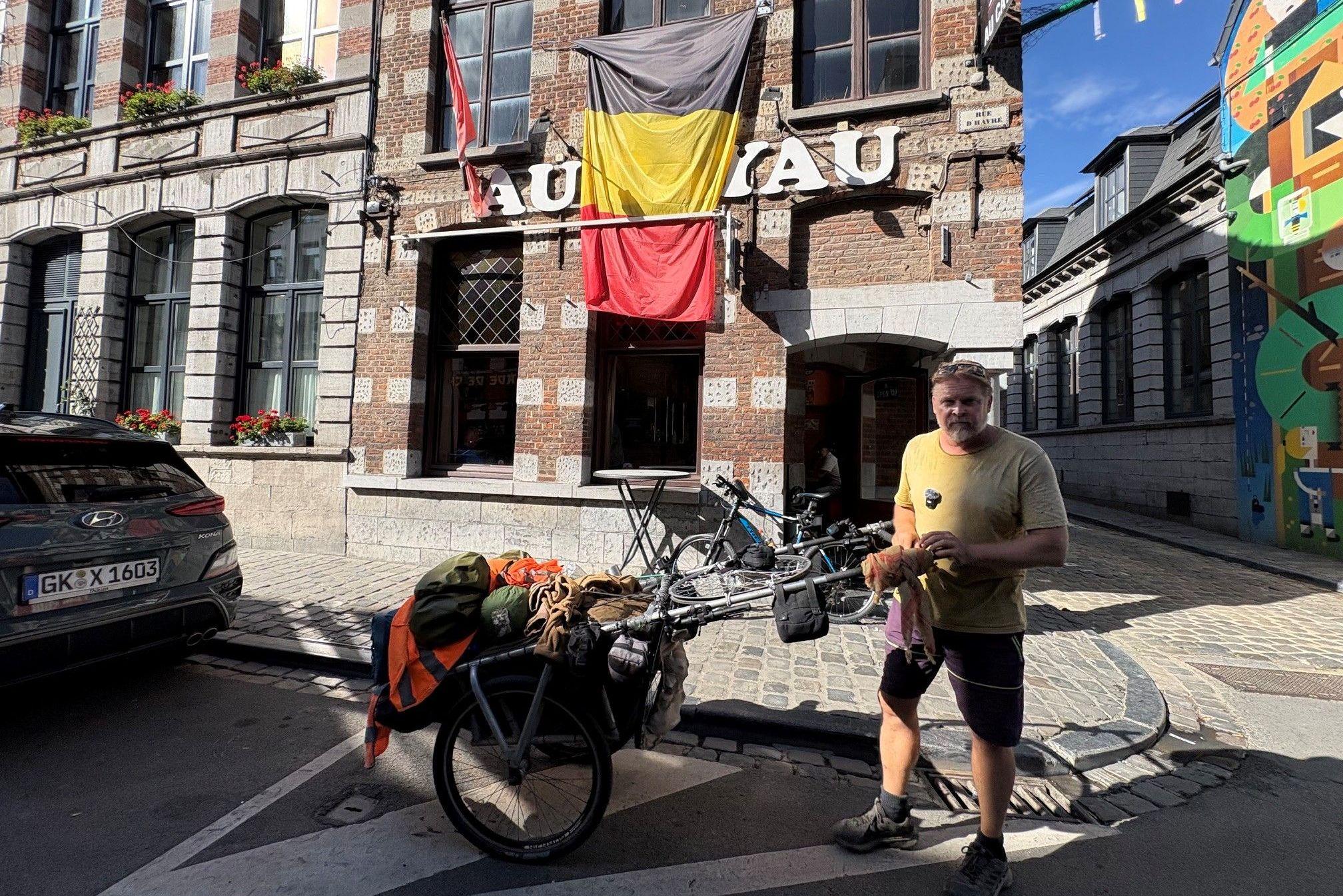 Un hombre de unos cincuenta y tantos años, con una camiseta caqui y pantalones cortos oscuros, posa en la calle junto a un carrito cargado de equipo y bolsas de camping. Una bandera belga cuelga de un edificio detrás de él.