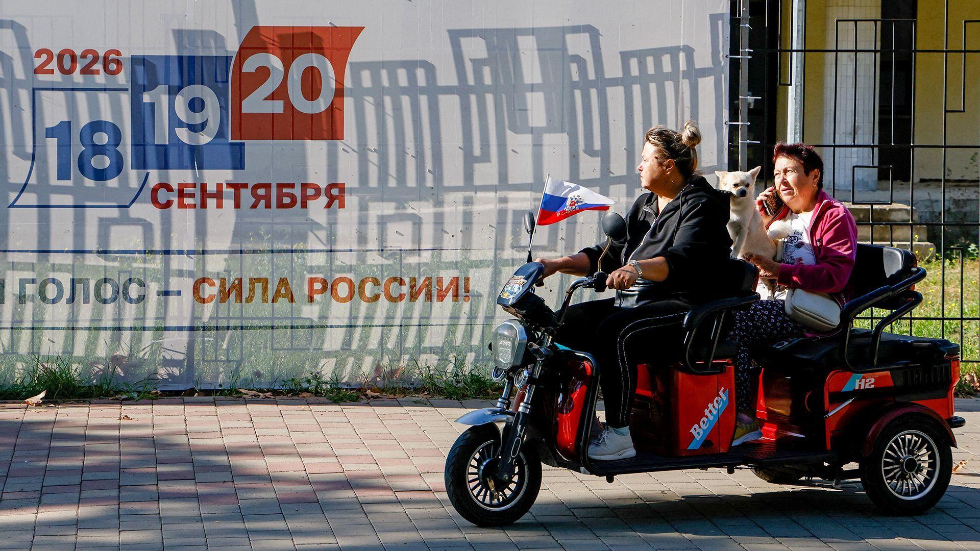 Dos personas circulan en una motocicleta eléctrica por una calle frente a una valla publicitaria que anuncia las próximas elecciones, en Crimea.