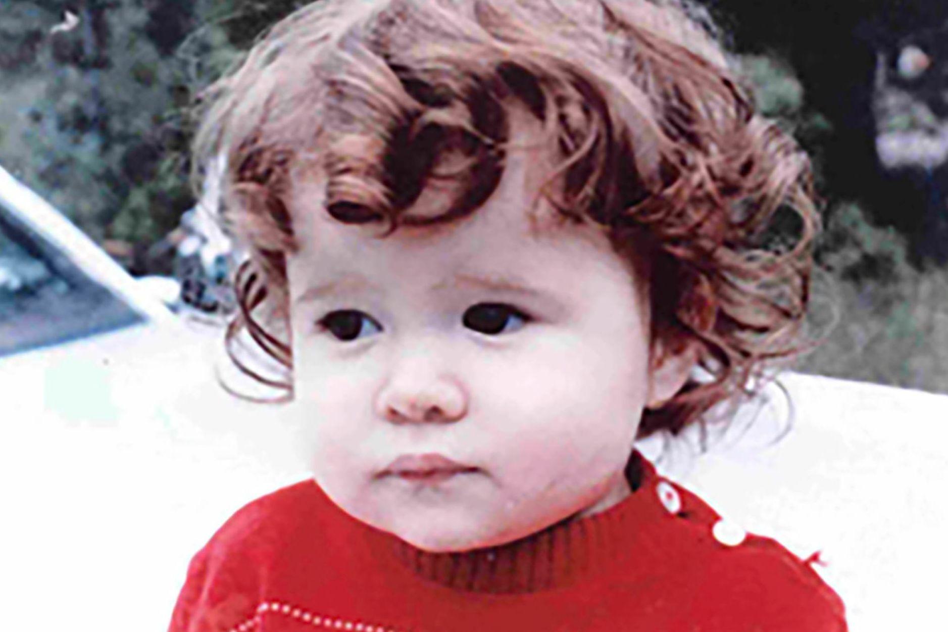 Christa Pike de niña. Tiene el pelo castaño, corto y rizado, y lleva un jersey rojo. Al fondo parece verse parte de un vehículo blanco.