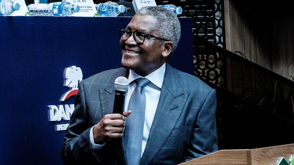 Aliko Dangote, vestido con traje, sonríe mientras gira la cabeza hacia la izquierda. Sostiene un micrófono en su mano derecha.