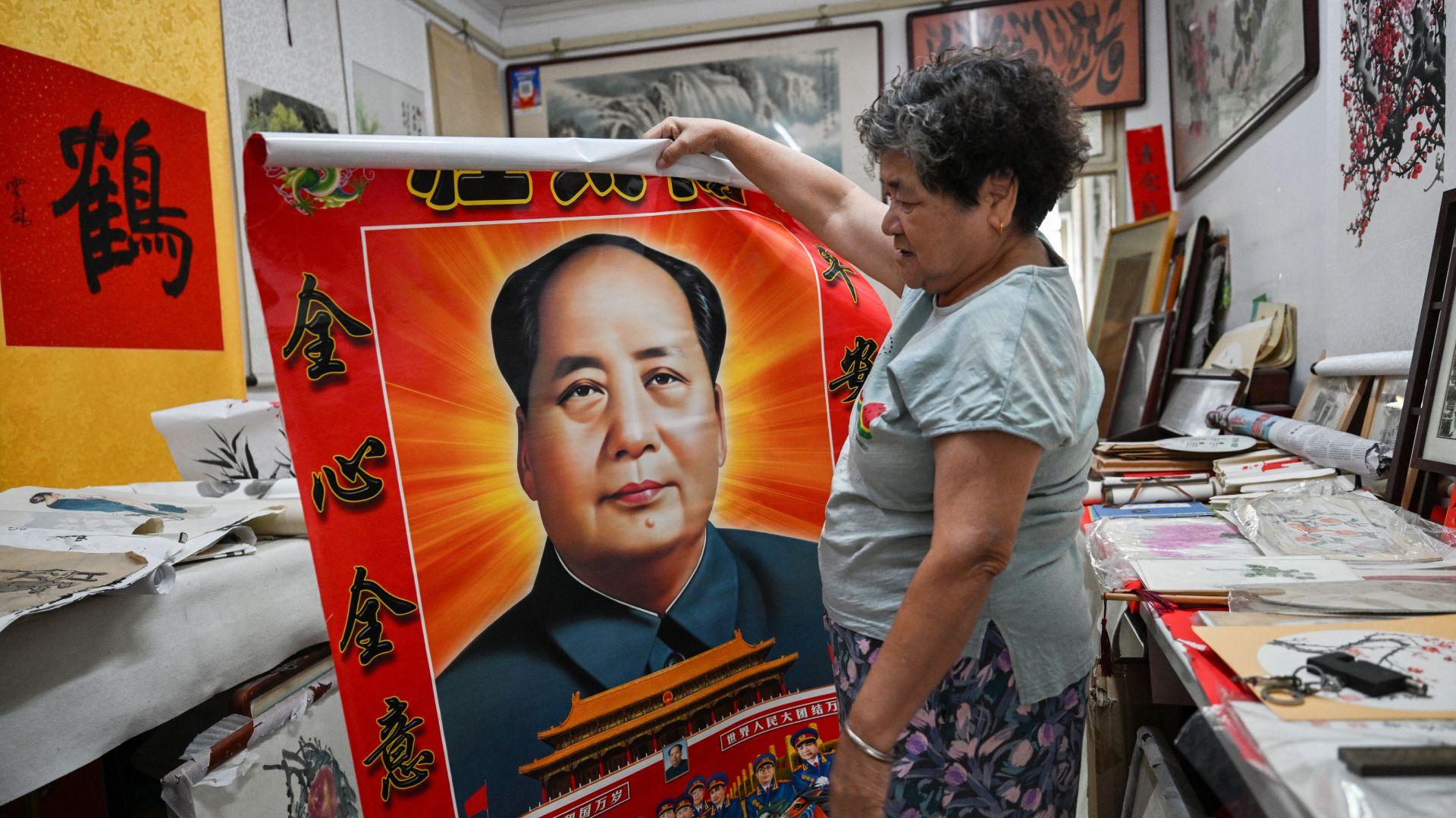 Una mujer despliega y sostiene un póster gigante de un Mao de gran tamaño, con luz que emana de su cabeza; debajo se puede ver la Puerta de Tiananmen y personas con uniformes militares.