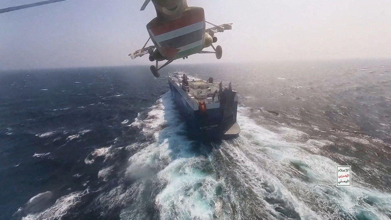 Se puede ver la parte inferior de un helicóptero hutí mientras se acerca por detrás al enorme buque de carga Galaxy Leader. El barco deja una estela inmensa a su paso mientras navega en mar abierto, sin que se vislumbre tierra firme.