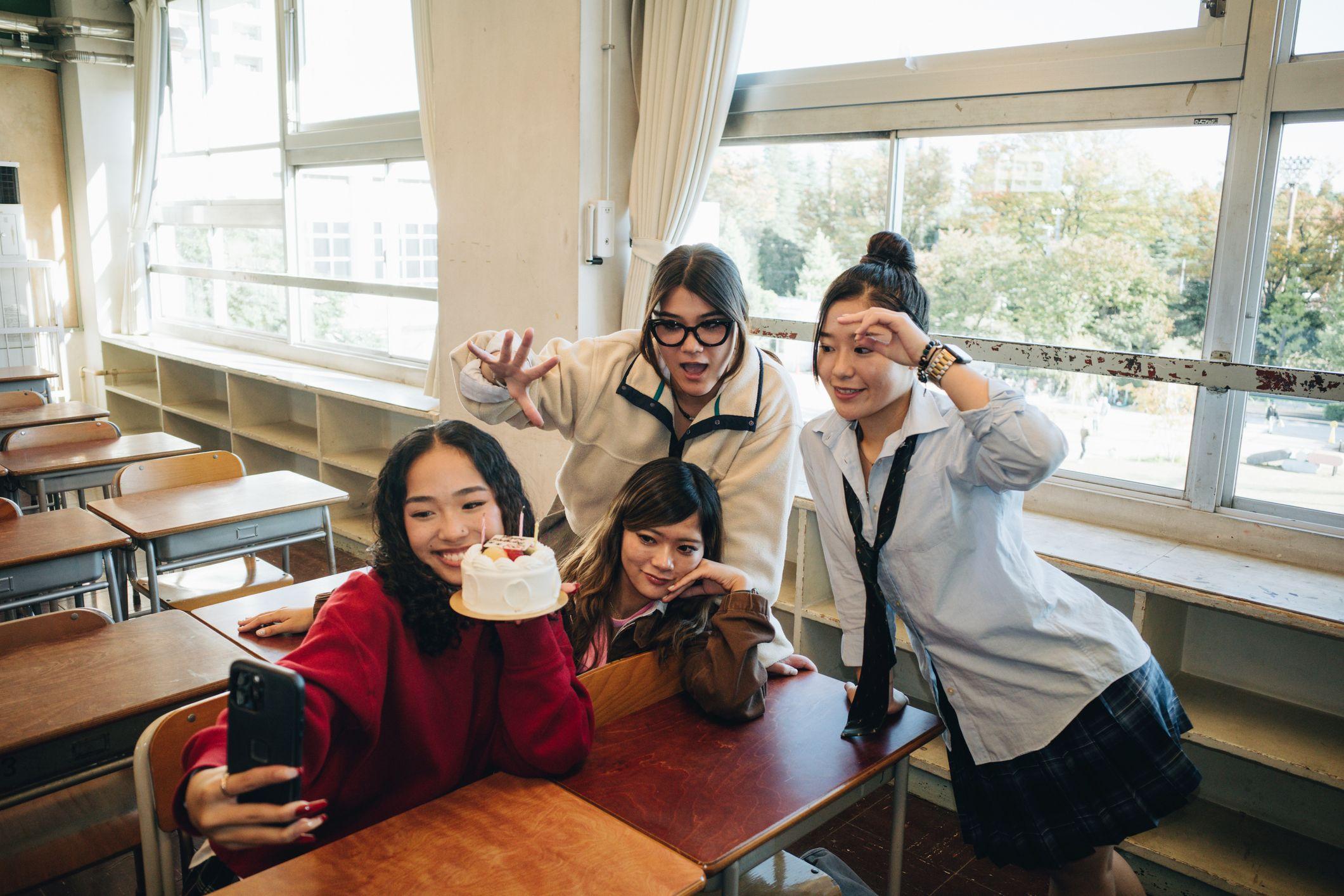 Cuatro chicas posan sonrientes mientras miran al teléfono que sujeta una de ellas. Está en un aula vacía. Dos están de pie y dos sentadas. 