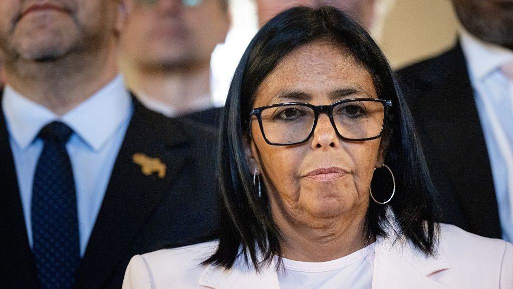 Delcy Rodríguez, en primer plano, vestida con traje de chaqueta claro. Lleva gafas y unos grandes aros en las orejas, Detrás de ella hay otras personas. 