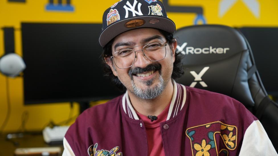 Hombre sonríe a la cámara. Lleva una vistosa chaqueta color carmesí y motivos de colores y flores. También lleva una gorra de los Yankees de Nueva York