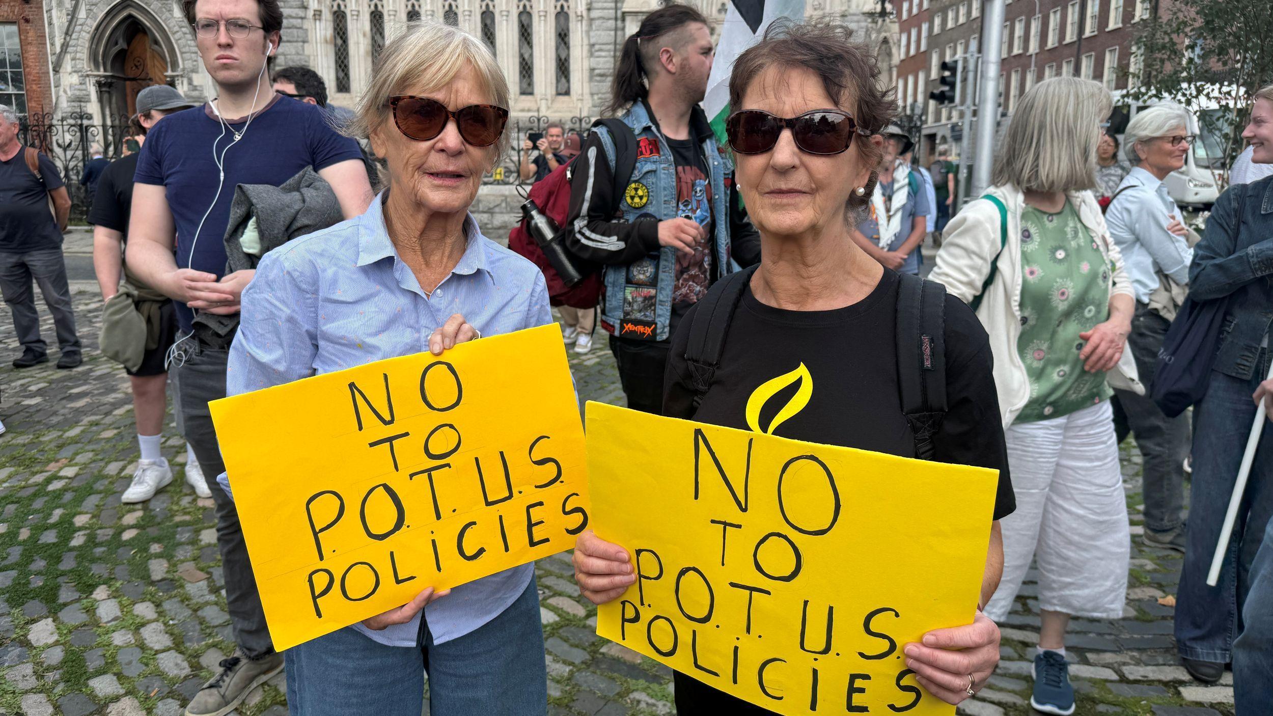 Una mujer de cabello rubio, gafas de sol, camisa azul claro y jeans está de pie a la izquierda con un cartel amarillo con letras negras que dice "No a las políticas del presidente". La mujer de la derecha tiene el cabello corto y oscuro, gafas de sol, una blusa negra y sostiene el mismo cartel.