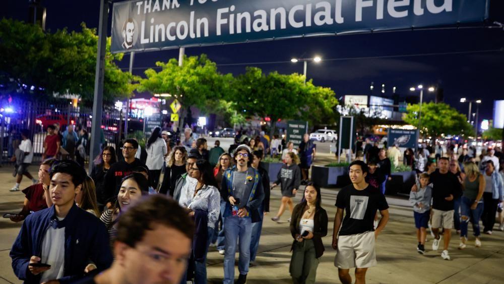 La gente camina al anochecer hacia el lugar donde se celebrará el concierto de la estrella del pop Ed Sheeran. Un letrero sobre ellos indica Lincoln Financial Field. 