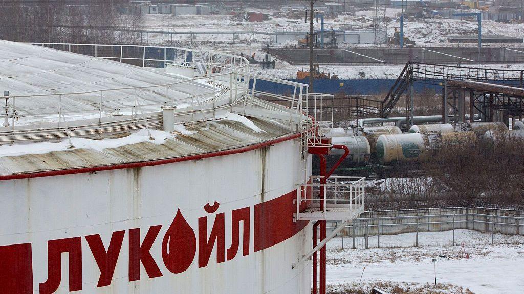 Un gran depósito blanco con el logo de la petrolera rusa Lukoil, en una instalación industrial cubierta de nieve, con vagones cisterna al fondo.
