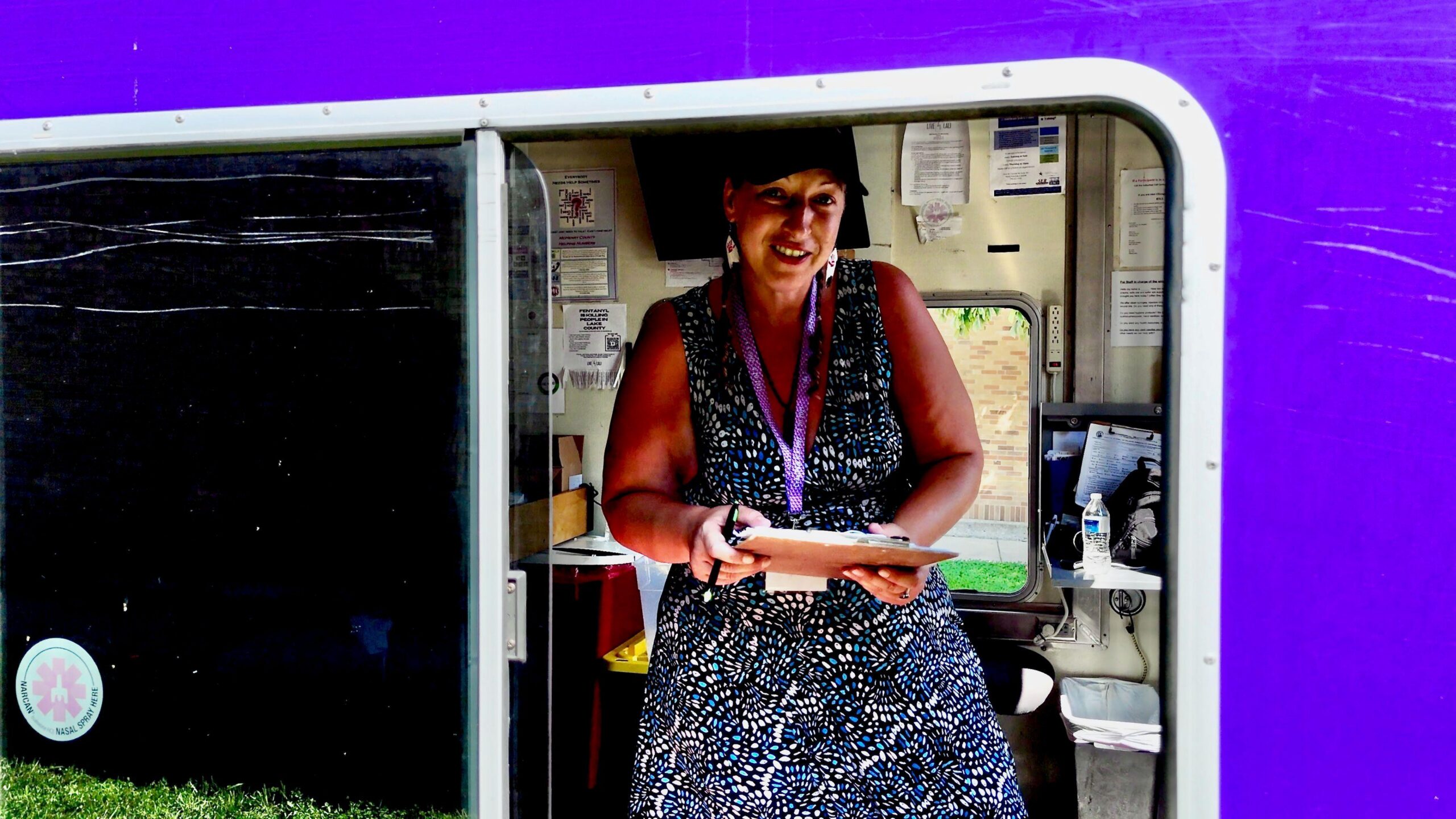 Karen aparece en la foto trabajando. Lleva un vestido azul con lunares blancos, una gorra de béisbol negra y un cordón morado. Sostiene un portapapeles y sonríe a la cámara