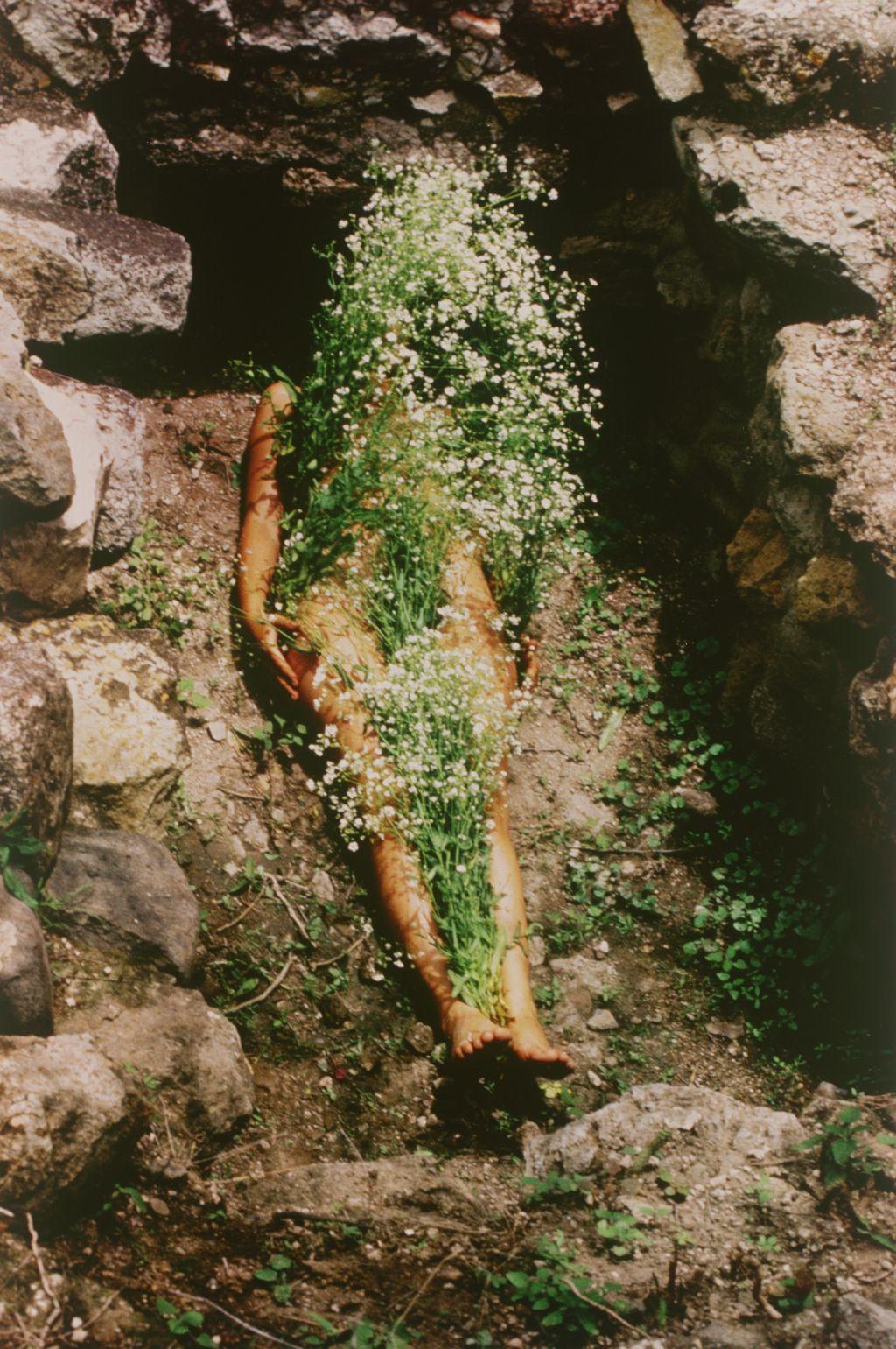 La artista acostada en una tumba de piedra con ramos de flores blancas cubriéndole el cuerpo desnudo