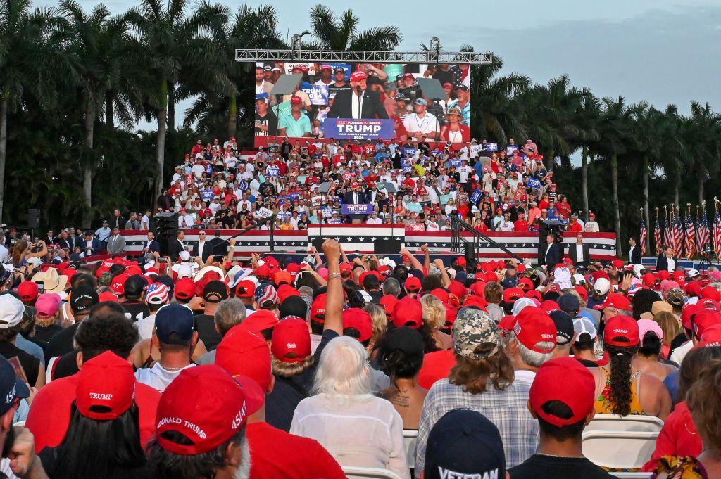 Donald Trump pronuncia un discurso en una tarima a lo lejos en un lugar al aire libre en Miami. De espaldas, mirándole, hay gran cantidad de personas con camisetas y gorras rojas.