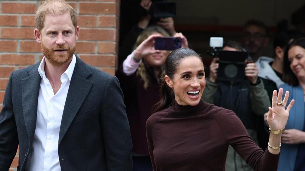 El príncipe Harry y Meghan Markle saludan a las cámaras que les apuntan. Meghan sonríe y levanta una mano. Harry tiene la boca entreabierta y usa un saco negro sobre una camisa blanca. 