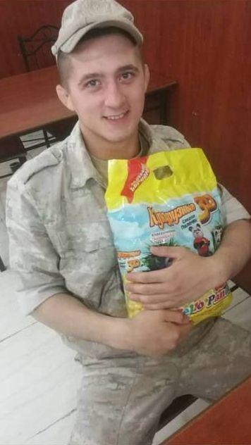 Artyom está sentado, vestido con ropa de camuflaje militar y una gorra. Sostiene un paquete amarillo. 