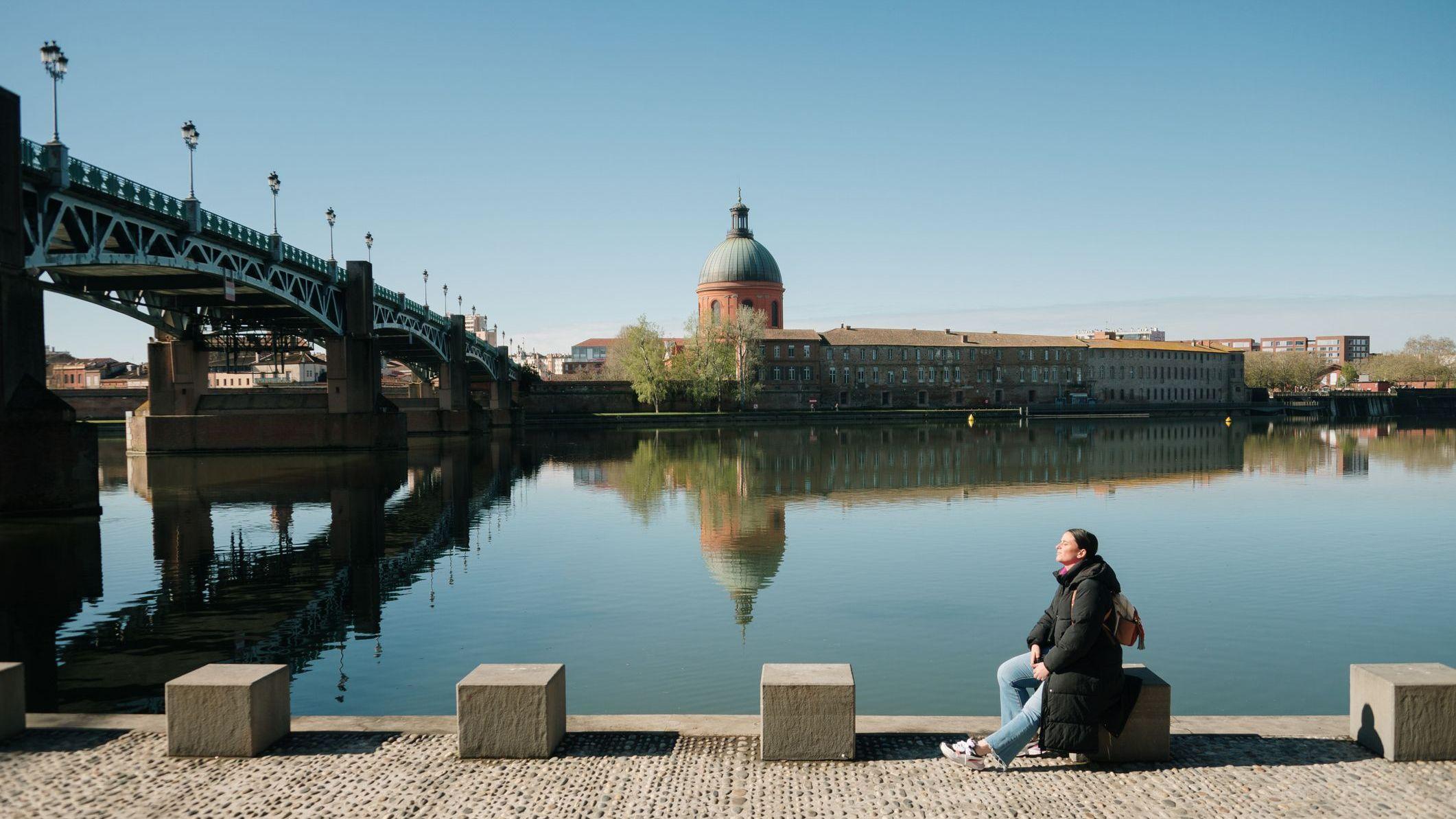 Una mujer que carga una mochila sentada a la orilla del río en Toulouse cerca de un puente; en el fondo, se ve un edificio con una cúpula.