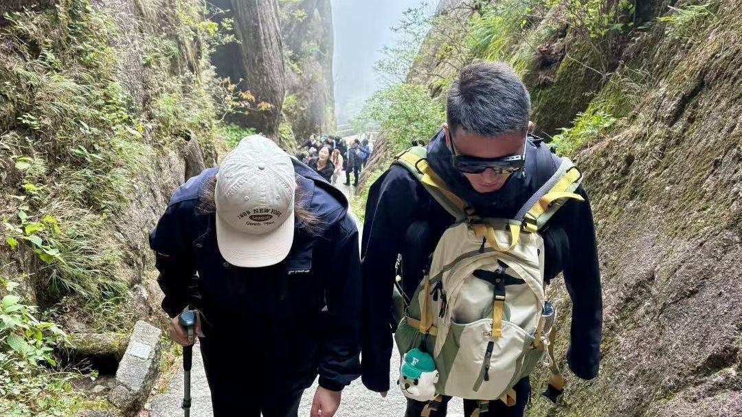 Fotografía tomada desde un ángulo elevado de dos excursionistas ascendiendo una empinada y brumosa escalera de montaña, enmarcada por acantilados rocosos.