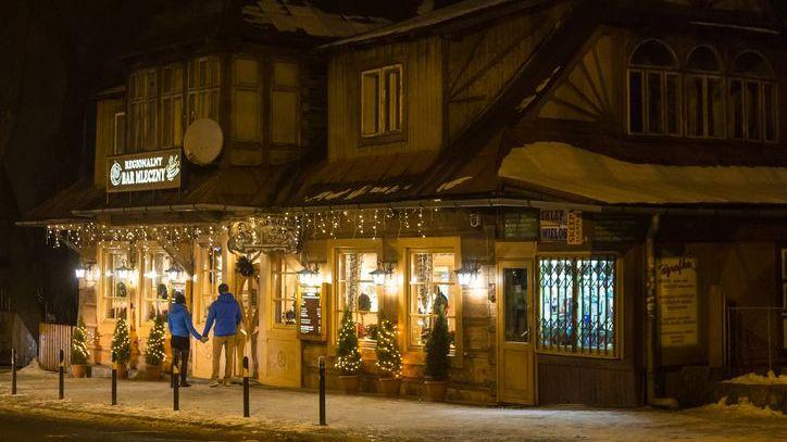 Un bar de leche en Polonia alumbrado con luces de navidad; en la puerta, un hombre y una mujer de espaldas, agarrados de la mano.