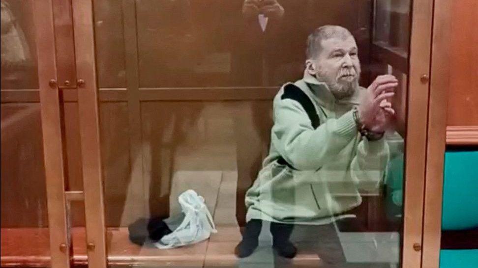 Stephen Hubbard, un hombre mayor que viste un jersey verde claro, permanece sentado esposado en una cabina de cristal en la sala del tribunal.