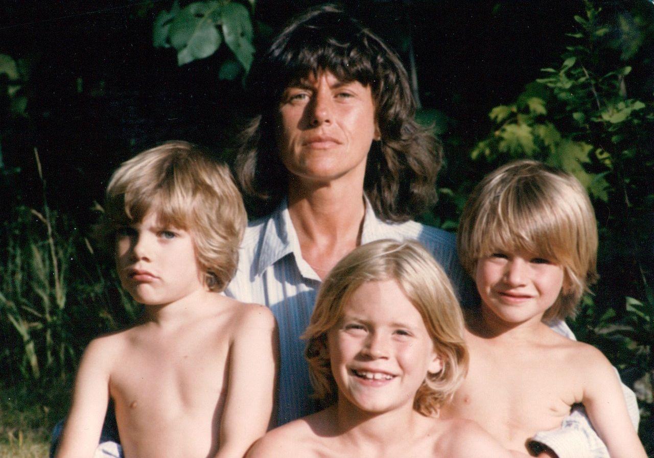 Una fotografía de retrato muestra a una mujer de cabello oscuro hasta los hombros sentada detrás de tres niños pequeños con el torso desnudo y cabello claro, todos muy juntos y mirando a la cámara. El grupo se encuentra al aire libre, en un entorno soleado y frondoso.