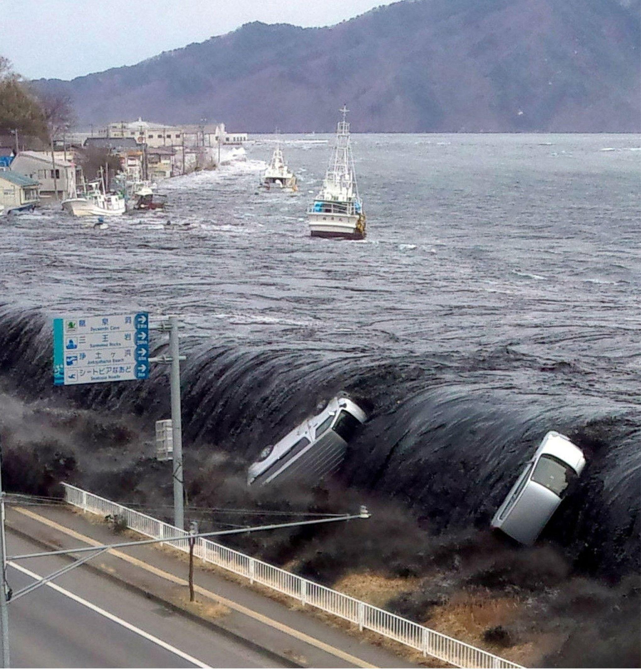 Una enorme y oscura ola de tsunami avanza tierra adentro a lo largo de una ciudad costera, arrastrando el agua de la inundación por carreteras y edificios, mientras que dos coches de color claro son levantados por la rápida corriente en primer plano. Al fondo, barcos de pesca permanecen en el puerto inundado.