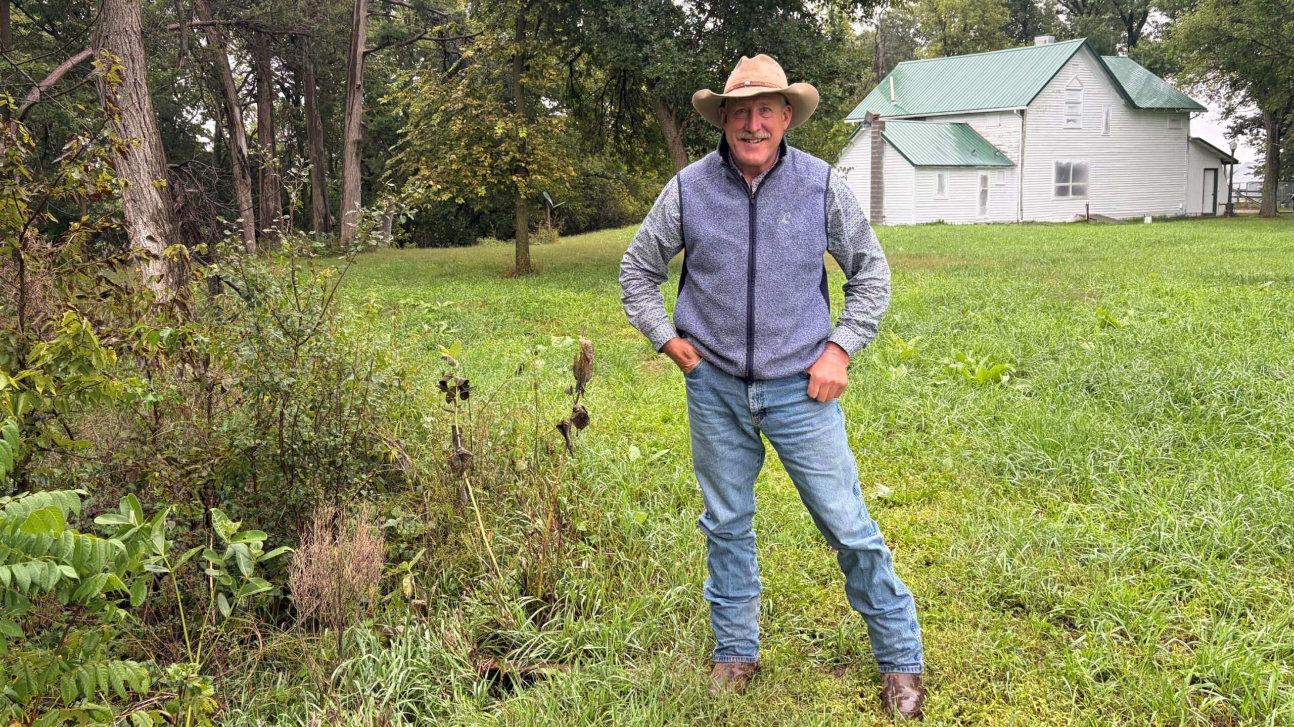 Jim Jenkins, con sombrero de vaquero, tejanos y chaleco, está de pie en su terreno.
