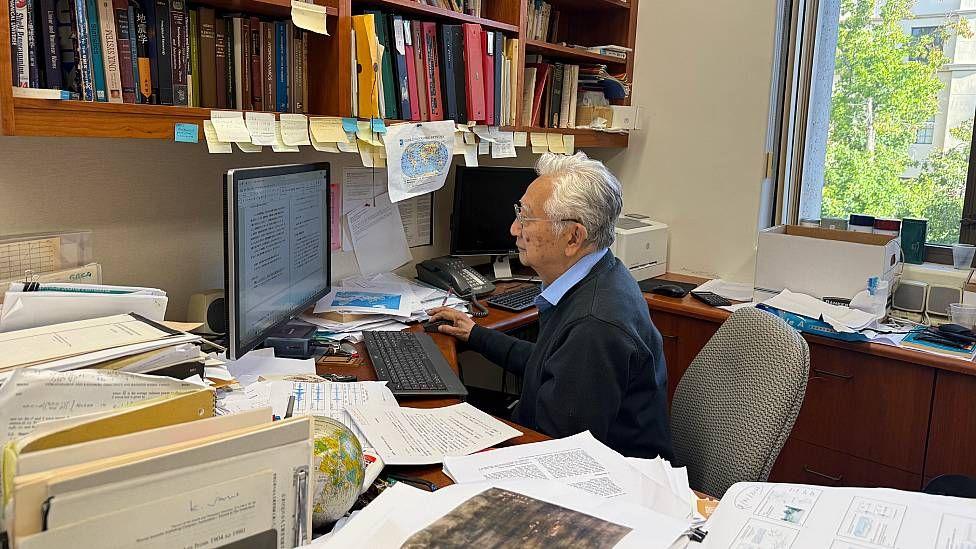 Hiroo Kanamori trabajando en su oficina en Caltech en setiembre de 2026. Está sentado frente al computador, su mesa está cubierta por documentos y sismogramas