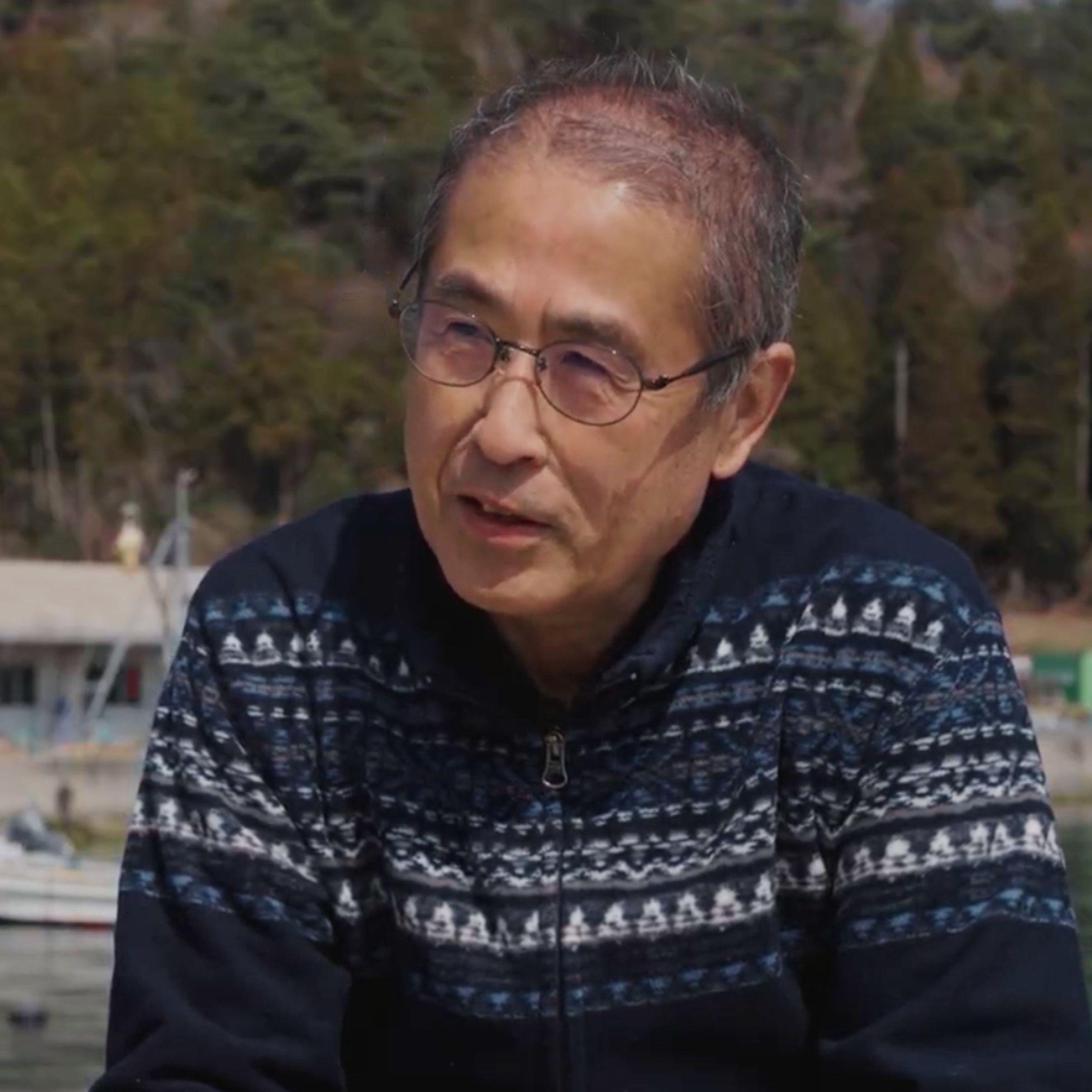 En la fotografía, un hombre (Yasuo) con gafas y un suéter azul oscuro con cremallera y estampado blanco aparece en un plano medio corto, sentado al aire libre y ligeramente de lado. Detrás de él se aprecia un paisaje costero, con agua, barcos borrosos y una orilla arbolada al fondo.