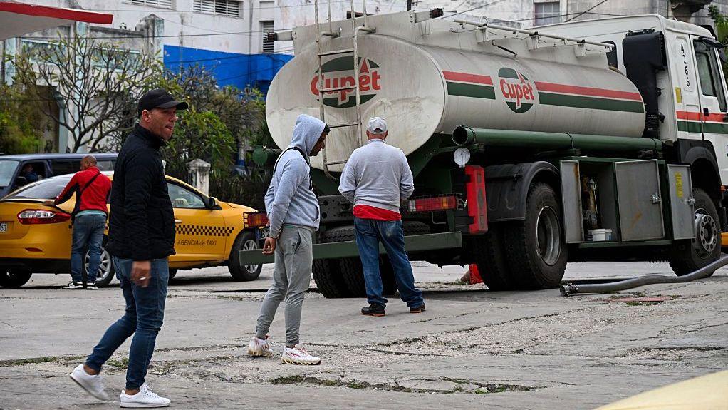 Un camión cisterna de la empresa estatal Cupet descarga combustible en una gasolinera de La Habana. Tres hombres esperan y en el fondo se ve un taxi amarillo.