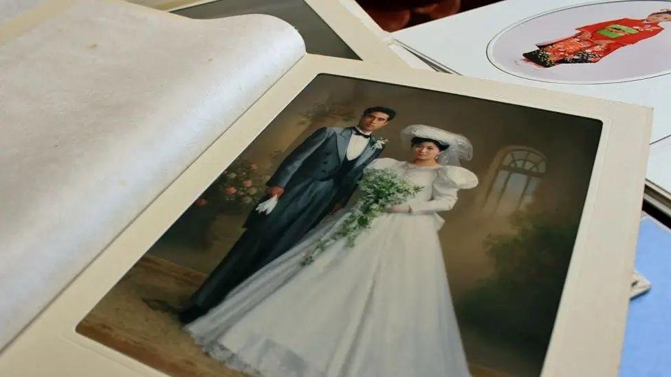Un álbum de bodas está abierto en una página que muestra a los novios. La novia (Yuko) lleva un vestido blanco largo con mangas abullonadas y un velo. El novio viste un traje gris formal y sostiene un par de guantes blancos. 