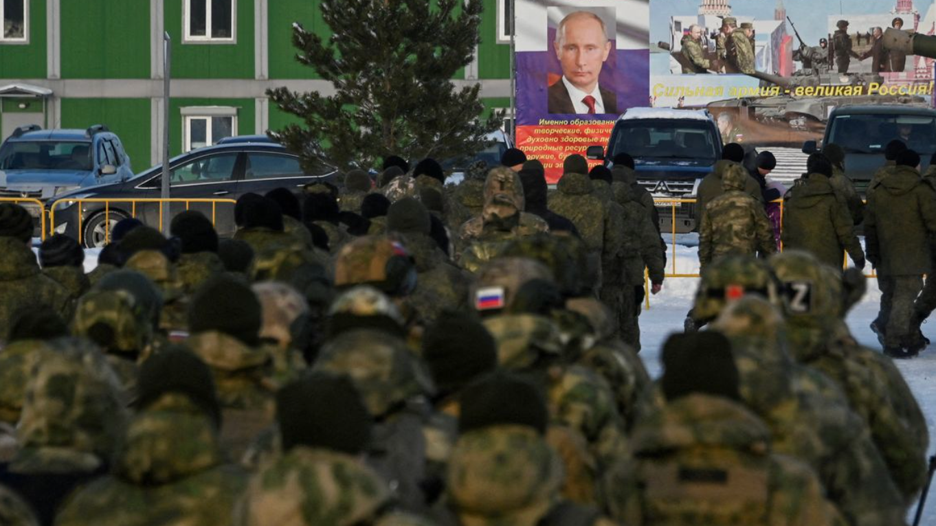 Las espaldas de un grupo de soldados vestidos con uniformes de camuflaje militar con la bandera rusa. Están frente a un cartel de Vladimir Putin.