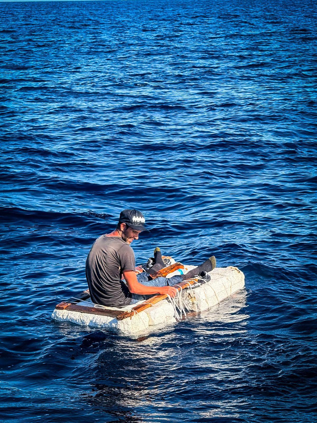 Un hombre sentado sobre una balsa improvisada que flota en el mar.