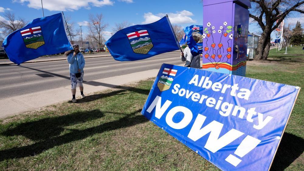Un hombre está de pie en la acera y ondea una bandera azul de Alberta. Sobre el césped, frente a él, hay un gran cartel del mismo color azul, con el escudo provincial, que dice: "¡Soberanía de Alberta ahora!"