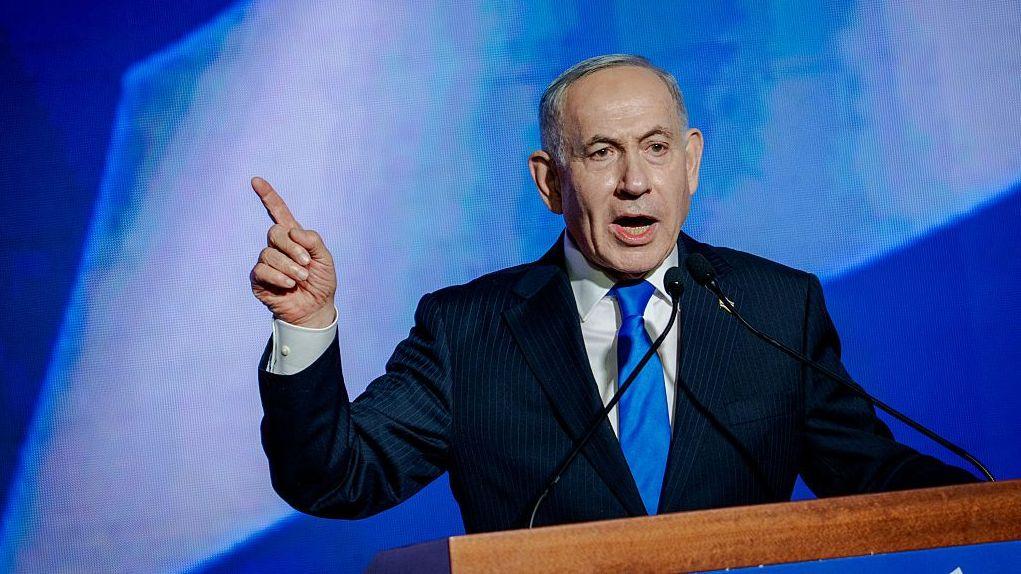 Se ve al primer ministro Benjamin Netanyahu hablando en un acto de campaña. Viste un traje azul con camisa blanca y corbata azul. El primer ministro tiene el dedo índice de la mano derecha apuntando hacia arriba.