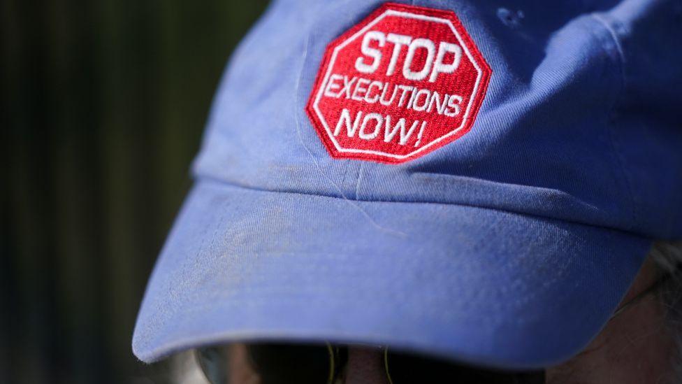 Gorra de béisbol azul con las palabras ¡ALTO A LAS EJECUCIONES AHORA! escritas en letras blancas sobre una insignia roja. No se ve el rostro de la persona que la lleva puesta, solo sus gafas de sol.