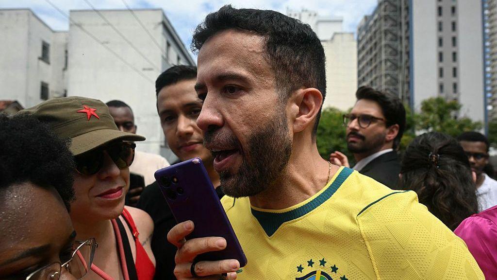Un hombre vestido con la camiseta amarilla de la selección de fútbol de Brasil habla por un celular. Esta rodeado de mucha gente. 
