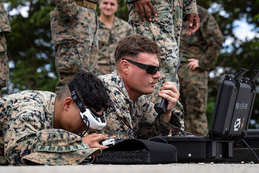 Dos militares de uniforme, tendidos en el suelo, operan un equipo de control remoto. El de la izquierda sujeta lo que parece ser un radiotransmisor con su mano izquierda. El de la derecha tiene los ojos cubiertos por lo que parece ser un equipo de visión de color blanco y parece manejar un mando de control. Tras ellos, de pie, se distiguen otros tres militares y la pierna de un cuarto que queda fuera de plano. 