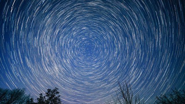 Fotografía de larga exposición del cielo nocturno, que muestra estelas de estrellas moviéndose en círculo alrededor de un punto central; el cielo es de un color azul medio y se pueden ver las copas de varios árboles en la parte inferior. 