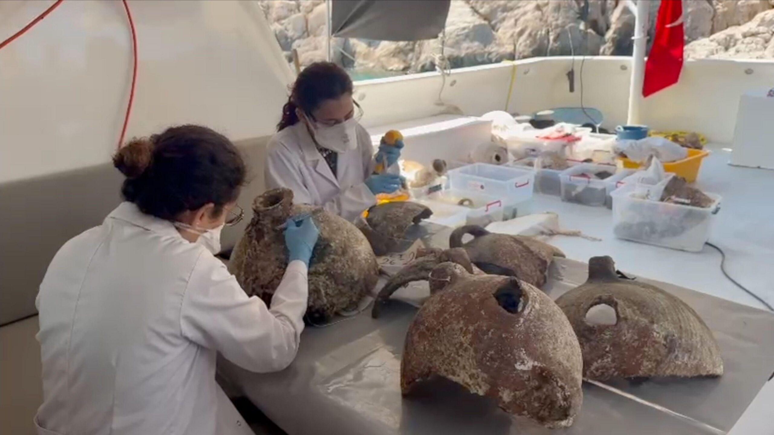 Dos personas con batas de laboratorio, guantes azules y mascarillas examinan algunas de las ánforas recuperadas. Están sentadas a una mesa en la parte trasera de un barco de buceo, a la sombra del techo. La persona de la izquierda, con el pelo recogido, observa una ánfora intacta. Con un bisturí, raspa suavemente la superficie cerca del cuello. Sobre la mesa hay otras cuatro ánforas parciales. Otra persona, sentada ligeramente detrás y a la izquierda, examina otro fragmento de cerámica. Al fondo, en la cubierta del barco, se ven seis cajas de plástico con más fragmentos de cerámica extraídos del fondo marino. Las rocas de la costa se divisan justo detrás de la popa, y parte de una bandera roja, sujeta a un mástil, cuelga en la esquina superior derecha.